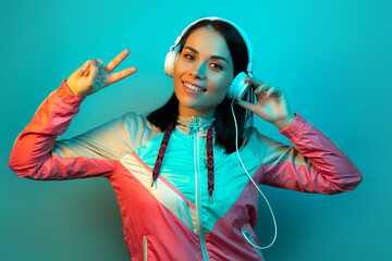 Smiling young woman in 90s style sport outfit with white headphonesshowing peace gesture in neon...