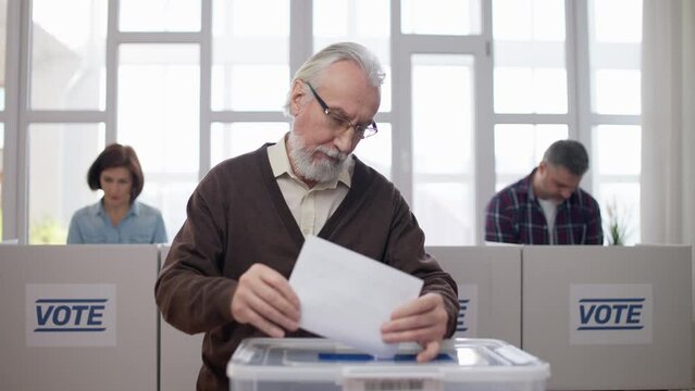 Senior man throwing voting ballot into box at polling station, citizens voting