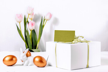 A box with an emblem label on a ribbon inside is not a big bento cake for the bright Easter holiday. Ceramic white rabbit with golden eggs on the background of spring delicate pink tulips.