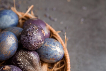 Purple, blue and golden eggs in a basket on a dark background. The purple hue trend of 2022 is very peri. Natural dye karkade tea. Top view. Easter card with a copy of the place for the text.