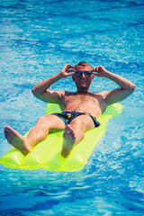 A young man bathes in the pool on a yellow inflatable mattress. Relax in the bright sun on vacation. Happy successful guy