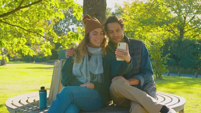 Couple sitting on bench in autumn park making video call on mobile phone - shot in slow motion 