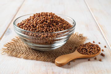 Close-up of raw wholegrain buckwheat groats in a glass bowl and wooden spoon over white wooden table. Ingredient for cooking porridge. Vegetarian food and gluten free diet concepts.