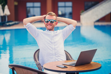 Successful handsome male businessman in sunglasses works at a laptop sitting near the pool. Remote work. Freelancer