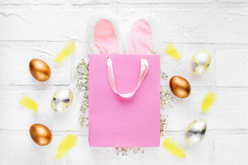A pink gift bag with white fluffy rabbit ears and marble Easter eggs with a sprig of hygrophila and yellow feathers. White brick wall. The concept of sales and discounts for Easter, delicate spring.