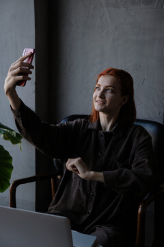 A red-haired woman takes pictures of herself on the phone while sitting in a cafe.