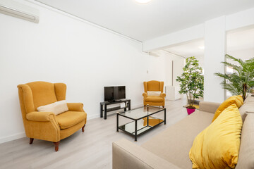 Living room with light brown 3 seater sofa, dark yellow cushions and matching wing chair with TV on black table, metal coffee table, air conditioning and light wood flooring