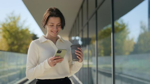 Positive caucasian business woman holding cup of coffee-to-go and using modern smartphone while standing near glass office building. Lifestyles, career and technology concept.