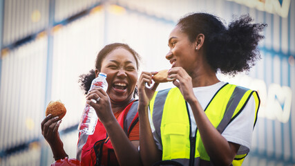 smile two African american woman foreman worker  or woman maintenance engineer in reflective vest safety jacket sits down on old truck relaxing, eats bread snacks and water during brunch break