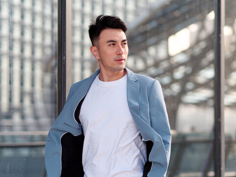 Portrait of handsome Chinese young man in light blue suit and white undershirt walking and looking away with modern city buildings background in sunny day, front view of confident businessman.