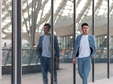Portrait of handsome Chinese young man in light blue suit and white undershirt walking and looking away with his reflections in glass wall background in sunny day, front view of confident businessman.