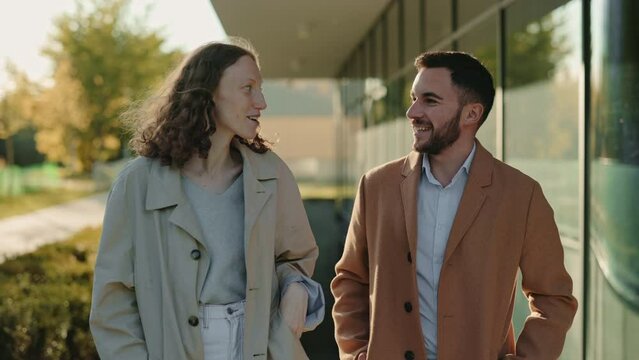 Positive young man and woman in smart casual clothes walking and talking near modern business center. Conversation of two coworkers. Business and lifestyles concept.