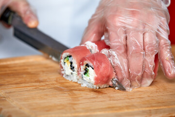 Chef cuts sushi rolls with red salmon fish with a knife on a wooden board. Chef in transparent...