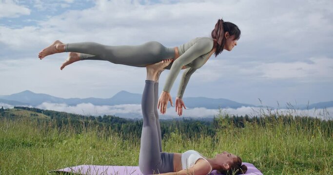 Two caucasian women doing balance acro yoga pose during summer time outdoors. Sporty ladies in activewear spending free time for workout on fresh air.