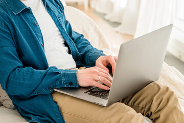 Hands a young man in a blue shirt using a laptop on his lap while sitting on the couch. Cozy conditions for remote work