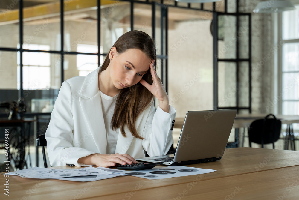 Wall mural stylish young female accountant looking through finances, using a laptop and a calculator, experienc