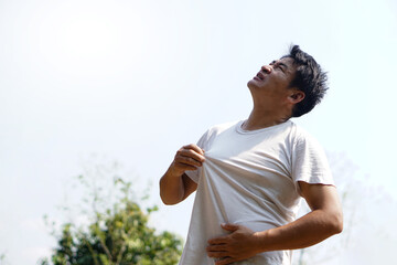 Asian middle-aged man feels hot, stands outdoor in sunny day. Concept : hot weather in summer, high temperature. Global warming. Heat wave. Uncomfortable feeling.     