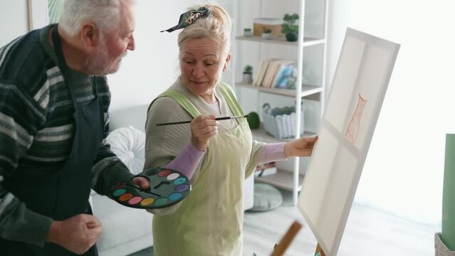 Cheerful old man giving painting lesson to elder female person attending art class program.