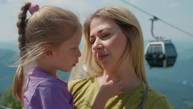 Attentive blonde mother holds little daughter in arms listening to story against modern poma lift and foggy mountains at resort closeup slow motion