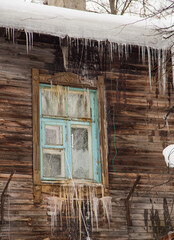 Glass transparent icicles hang on the edge of the roof and the window. Against the background of the wooden wall of the old house. Large cascades, even beautiful rows. Cloudy winter day, soft light.