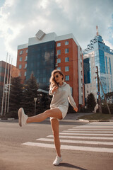 teen walk. young blonde curly girl in sports gray costume is walking with funny jump and smiling happy in sunglasses on the street buildings background. sport lifestyle concept, free space