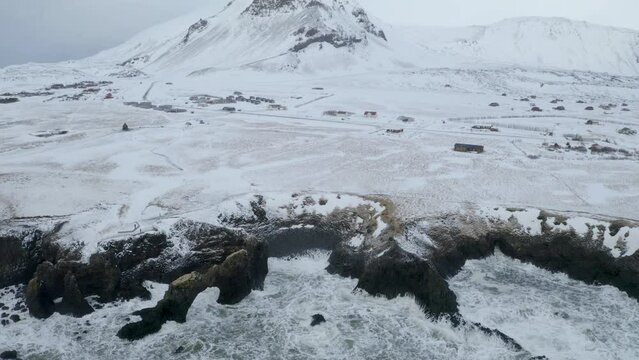 Aerial tilt down shot of snowy Sn&aelig;fellsj&ouml;kull Volcano and waves of ocean with steep dark cliffs in foreground - Visiting Iceland by Winter