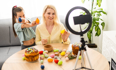 Cute little girl and beautiful young mom sitting together at kitchen table using smart phone for online at easter