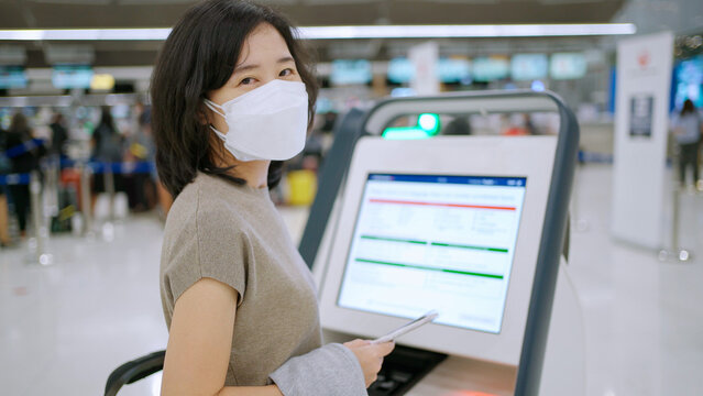 Woman Wear Mask Using Self Service Check-in Kiosk At The International Airport Terminal. Touchscreen And See Information On Screen. Scan Passport To Record Personal Data. New Normal Travel