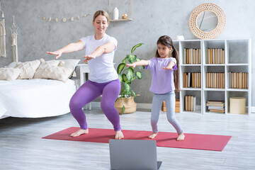 Young mother and little daughter exercising online on fitness mat at home with laptop