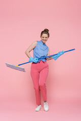 full length of cheerful young woman in rubber gloves imitating play on guitar with mop on pink.