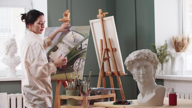 A woman artist puts undone painting on an easel and continue drawing