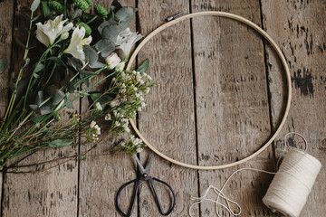 Making stylish spring wreath with beautiful fresh flowers. Wooden hoop, thread, scissors, and flowers on rustic wooden background. Modern floral arrangement and handmade decor.