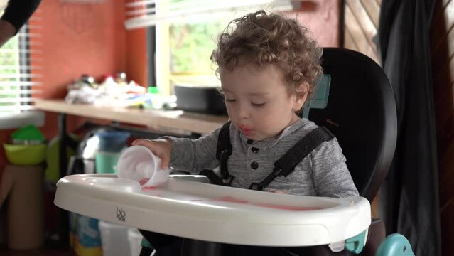 little caucasian latino baby sitting and sipping a drink in his dining chair at home