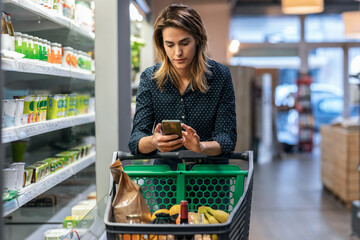 Modern woman using smartphone with trolley cart while walking and taking products from shelf in a...