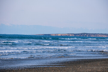 Stormy sea waves hit a sandy beach. Snow capped mountains appear in the distance.