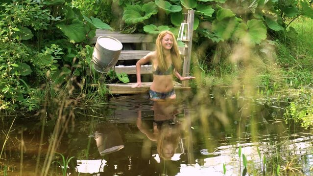 A slender young woman swims in a pond among reeds and burdocks. Rural scene.