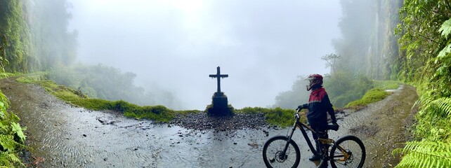 Panoramic view of mountain biker near grave on edge of foggy mysterious Death Road, Bolivia - obrazy, fototapety, plakaty