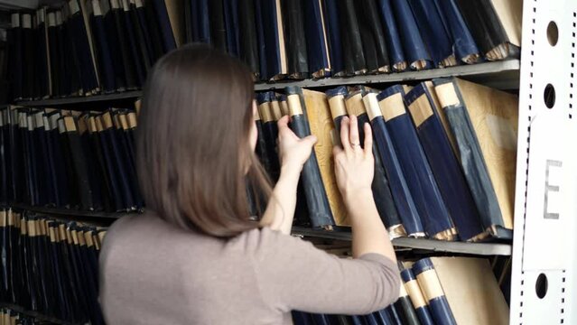 Young woman on a shelf in the archives of the library is looking for the right patent.