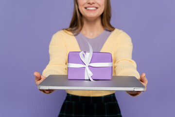 Obraz premium cropped view of happy young woman holding laptop with gift box isolated on purple.