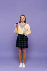 full length of happy young woman holding laptop and paper cup on purple.