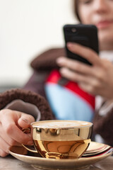 Young woman taking a photo of coffee with her smart phone. Girs face is not seen in photo. Focus is on the golden coffee cup. Girl is blur. Vertical photo.