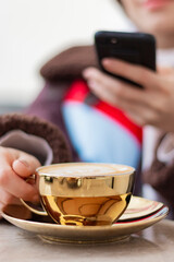 Young woman taking a photo of coffee with her smart phone. Girs face is not seen in photo. Focus is on the golden coffee cup. Girl is blur. Vertical photo.