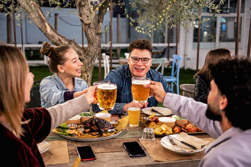 Happy friends dining and tasting beer glasses together in garden in barbecue dinner party - Smiling...
