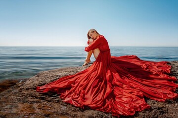 Beautiful sensual woman in a flying red dress and long hair, sitting on a rock above the beautiful...