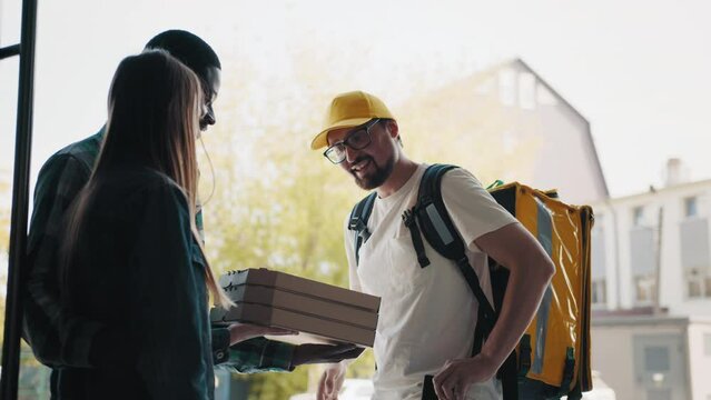 Fast pizza delivery. View of a young courier carrying boxes of pizza. Handsome positive man, takes orders, pays by credit card. Multiracial people delivering food to home or office.