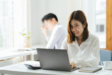 Attractive asian young confident businesswoman sitting at the office table with group of colleagues in the background, working on laptop computer