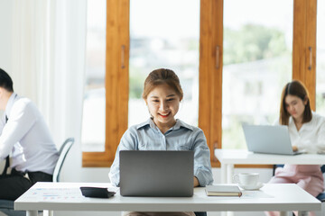 Smiling young asian businesswoman working on a laptop at her desk in a bright modern office with colleagues in the background