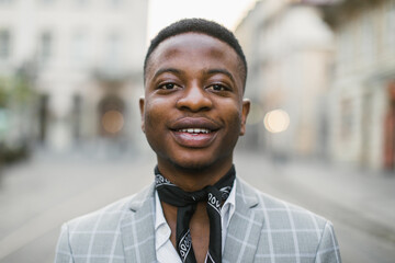 Portrait of handsome african american man in stylish suit and scarf smiling on camera while posing outdoors. Blur background of city street.