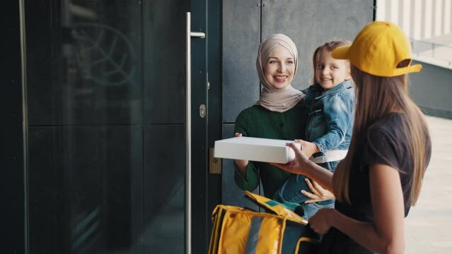 Parcel for a child. The courier girl brought the parcel to the family. A positive Muslim woman in a hijab with a child accepts her online order from a courier girl in a yellow cap and thermal backpack