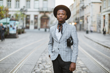 Positive african man in stylish suit and hat walking on city street with vintage photo camera in hands. Male tourist enjoying vacation at new places.